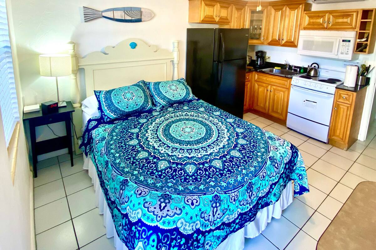 A bedroom with a bed, bedside table, lamp, and a view of a kitchen with a refrigerator, stove, and microwave.