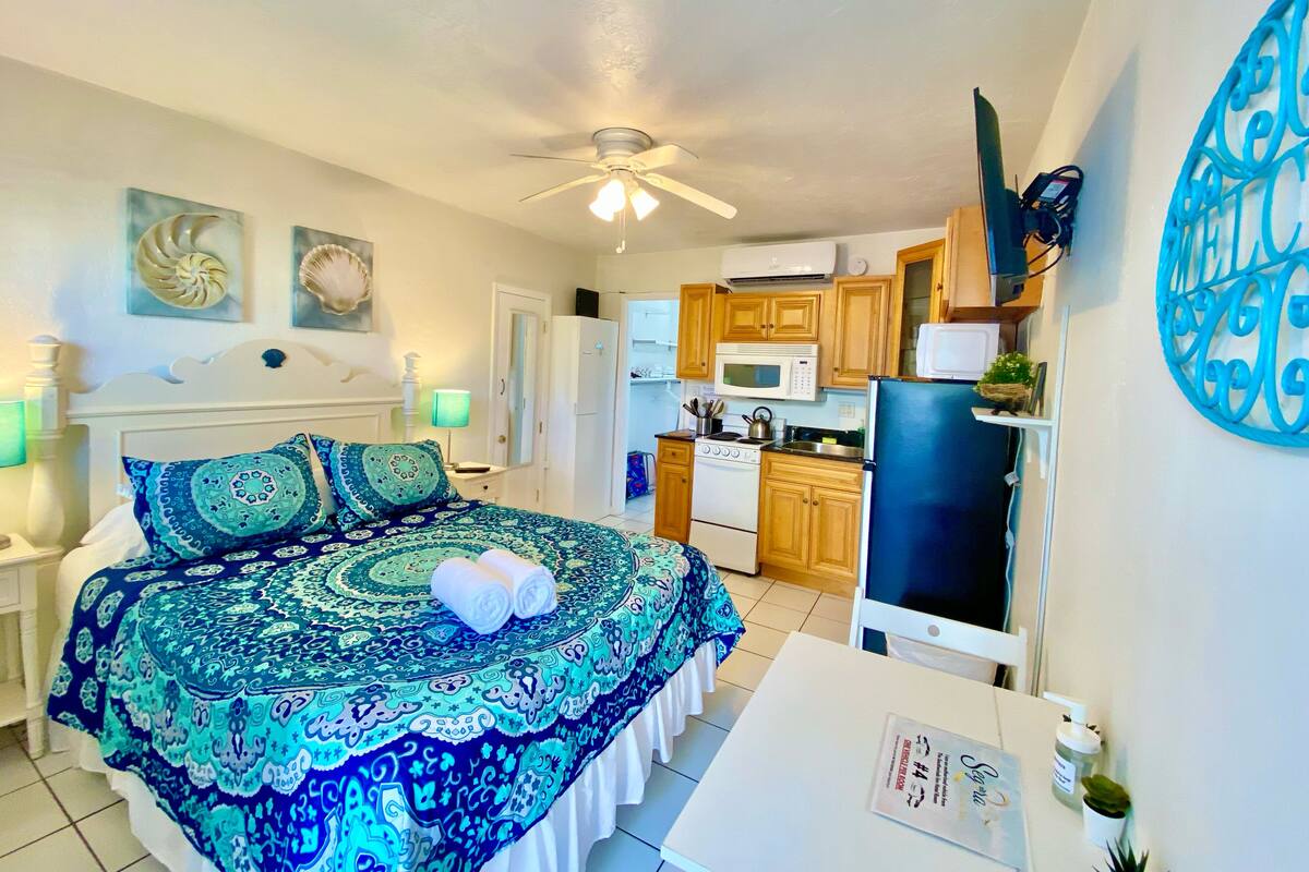 A bedroom with a large bed, a ceiling fan, and a view into the kitchen area.
