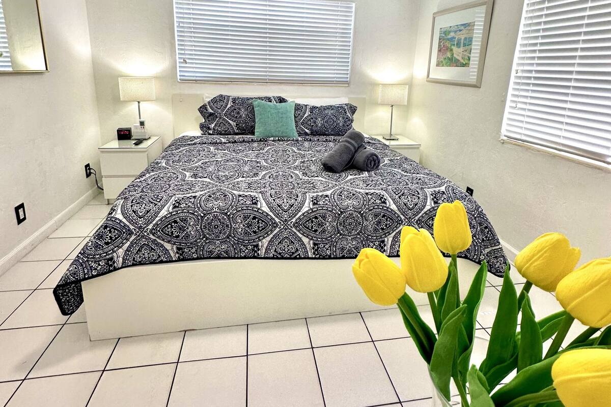 A bedroom with a bed, a ceiling fan, a nightstand, a lamp, and a window with blinds.
