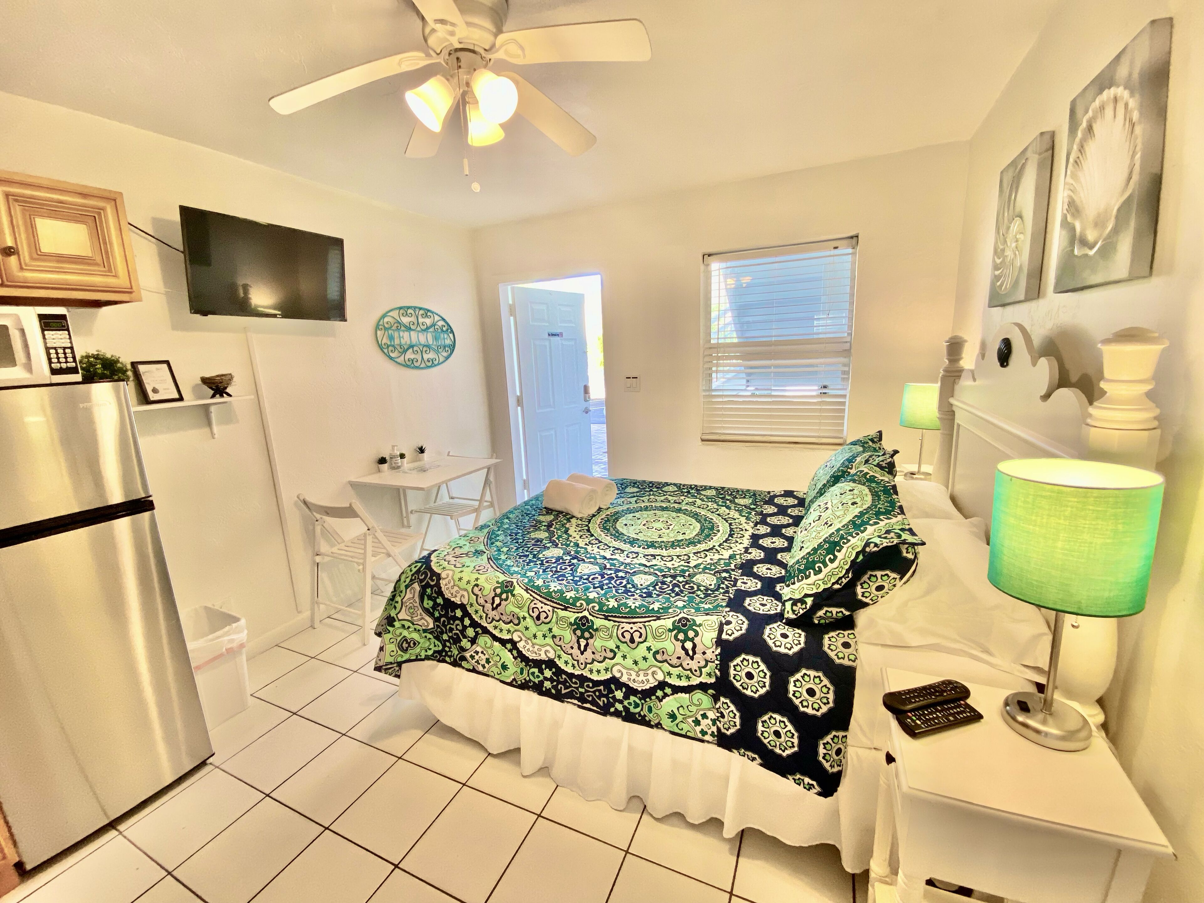 A compact hotel room with a bed, a desk, a chair, a refrigerator, a ceiling fan, and a window with blinds.