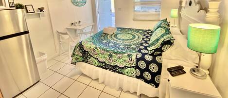 A compact hotel room with a bed, a desk, a chair, a refrigerator, a ceiling fan, and a window with blinds.