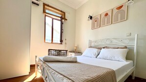 Un dormitorio con cama blanca, piso de madera, ventana con persianas, mesita de noche con lámpara y decoración en la pared.