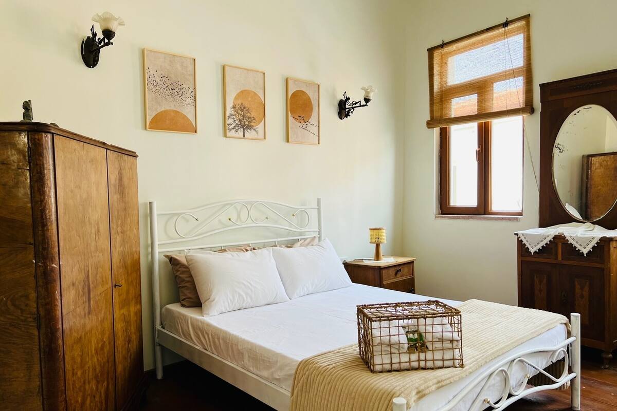 Quarto com estrutura de cama em metal branco, guarda-roupa de madeira, mesa de cabeceira com abajur e janela com persiana de bambu.