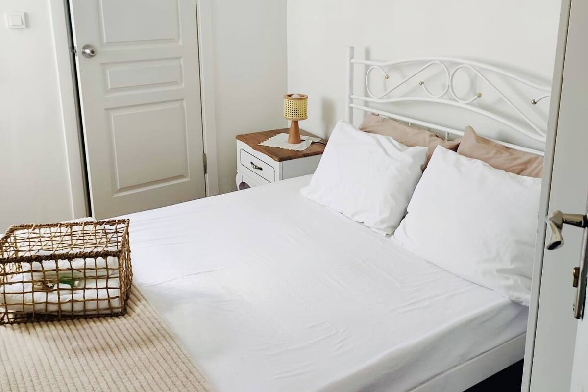 Cama bem arrumada com roupa de cama branca, mesa de cabeceira com abajur e um cesto de vime sobre a cama.