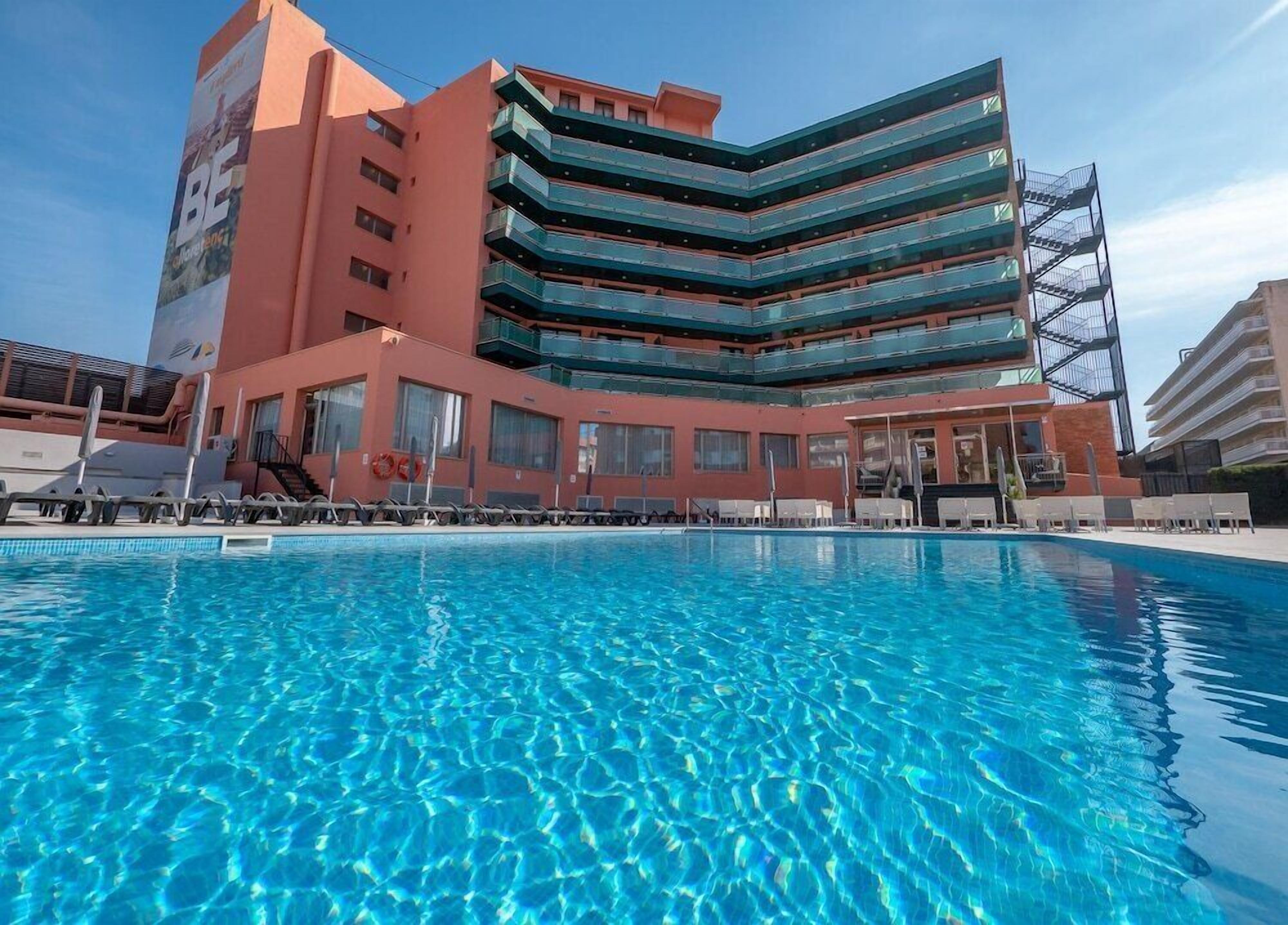 A modern hotel with a large pool, multiple balconies, and a clear blue sky.