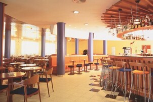 En rummelig hotellobby med flere siddeområder, en central bar og et stort vindue der giver naturligt lys.