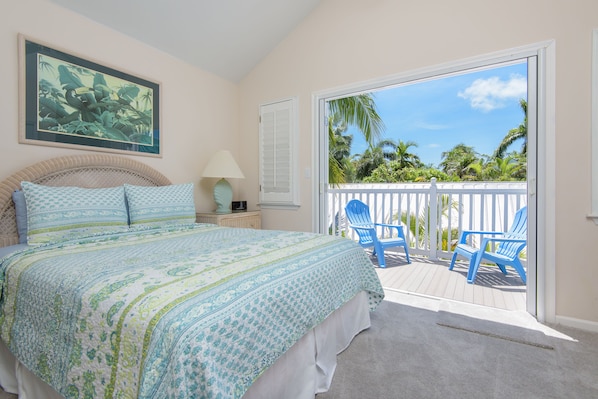 A bedroom with a large bed, a nightstand, a lamp, and a view of a balcony with blue chairs and a white fence.