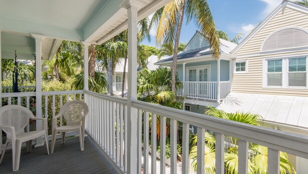 A covered porch with white railings, two white plastic chairs, and a view of a house with a blue roof and white siding. - Sago Palm at The Truman Annex in Key West (Key West)