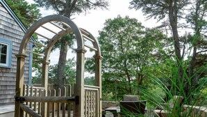A wooden gate with a metal frame, a stone pathway, and a barbecue grill in a garden setting. - Cozy Cockle Cove Retreat! (South chatham)