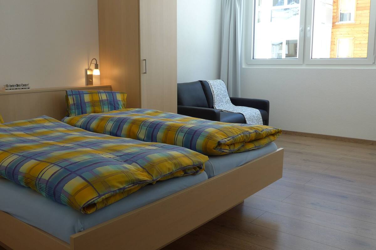 Un lit double avec une couette à carreaux, une table de chevet avec une lampe, une chaise et une fenêtre donnant sur des immeubles.