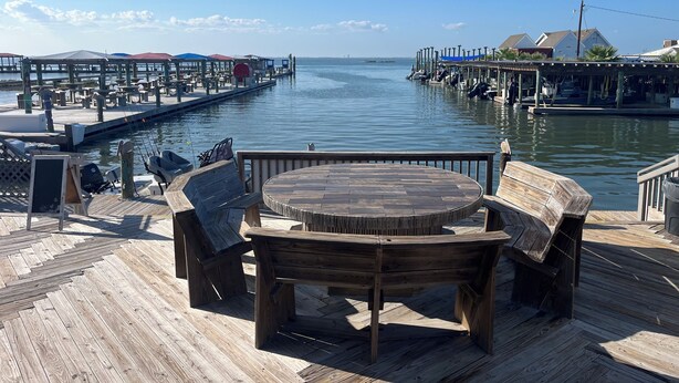 Una terraza de madera con una mesa redonda y bancos, con vistas a un puerto deportivo con barcos y un muelle.