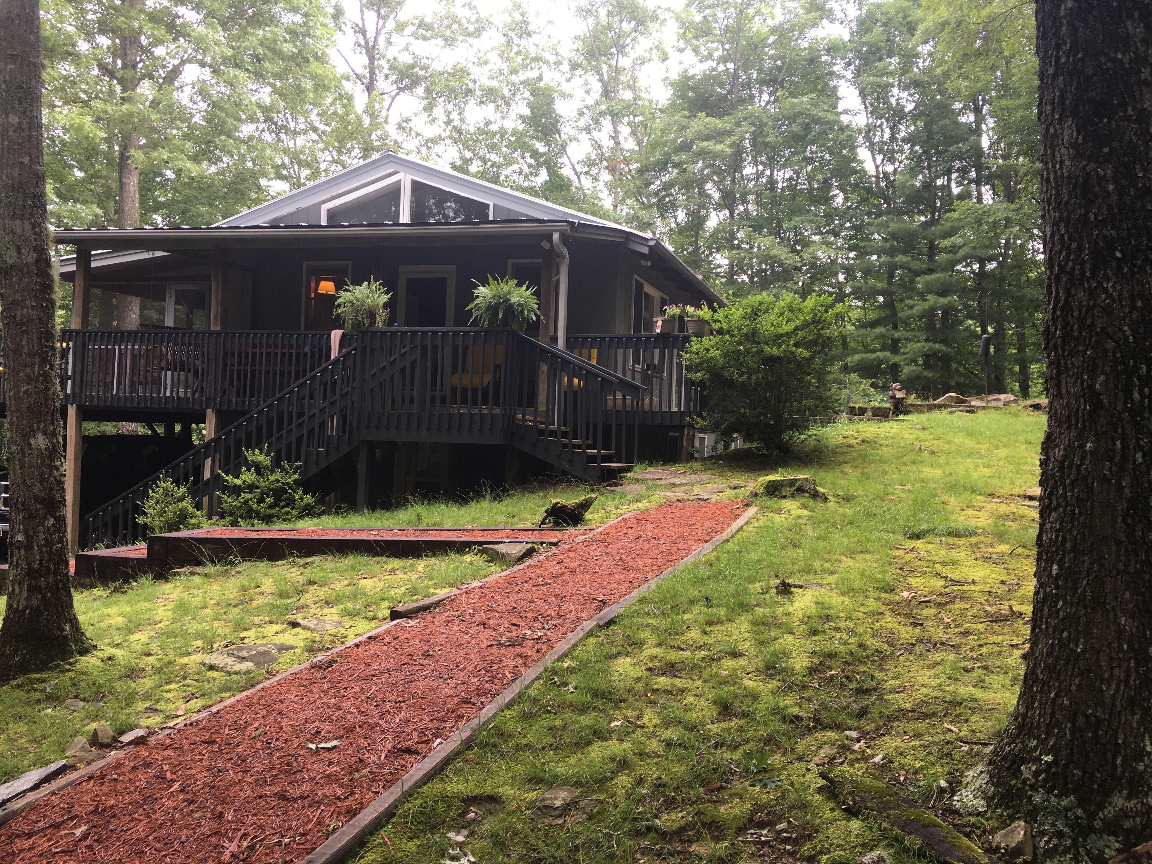 A two-story cabin with a wooden deck and a covered porch, surrounded by trees and a gravel path.