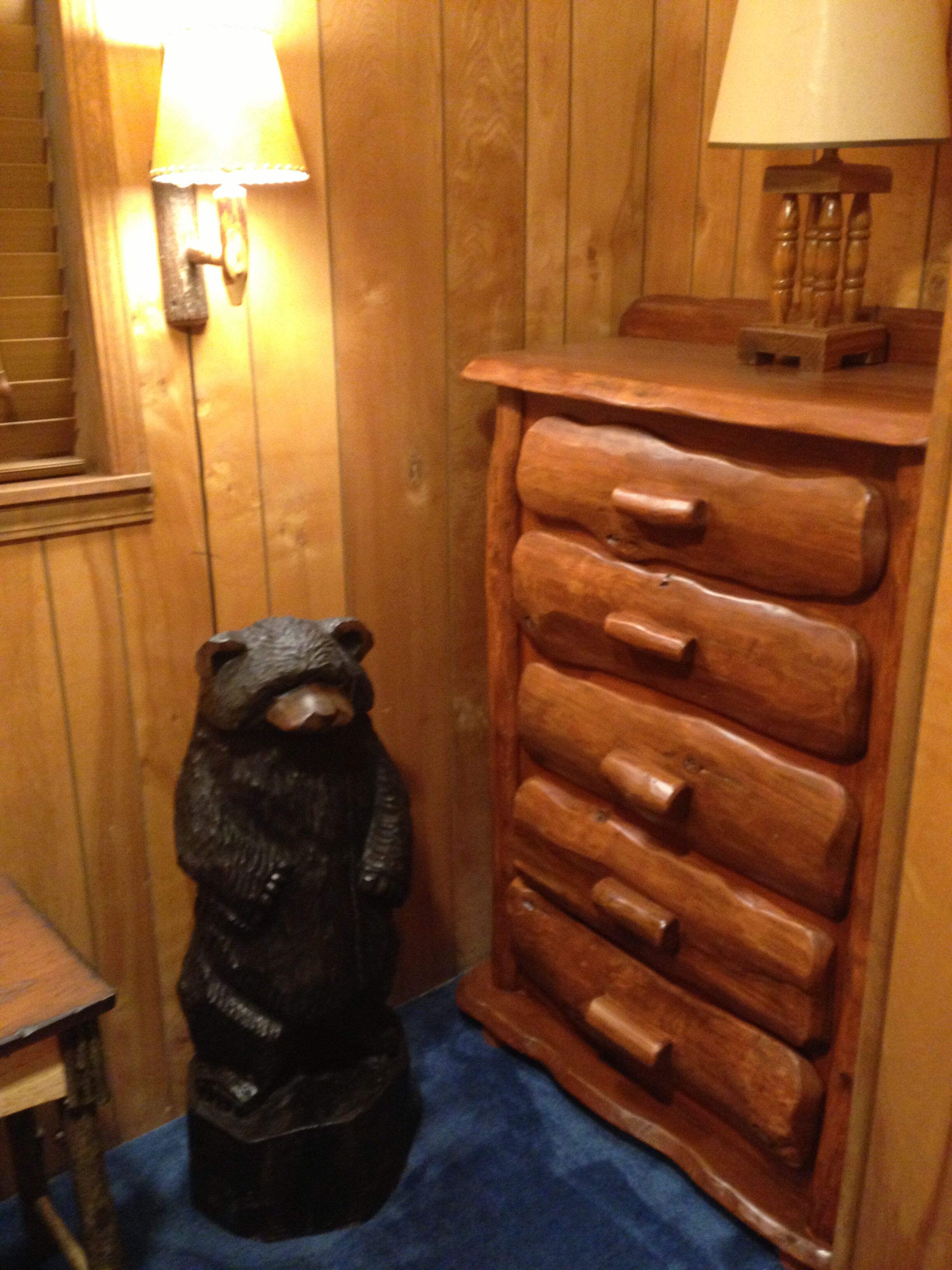 Une statue d’ours en bois dans un coin, une commode en bois avec des tiroirs et une lampe posée sur la commode.