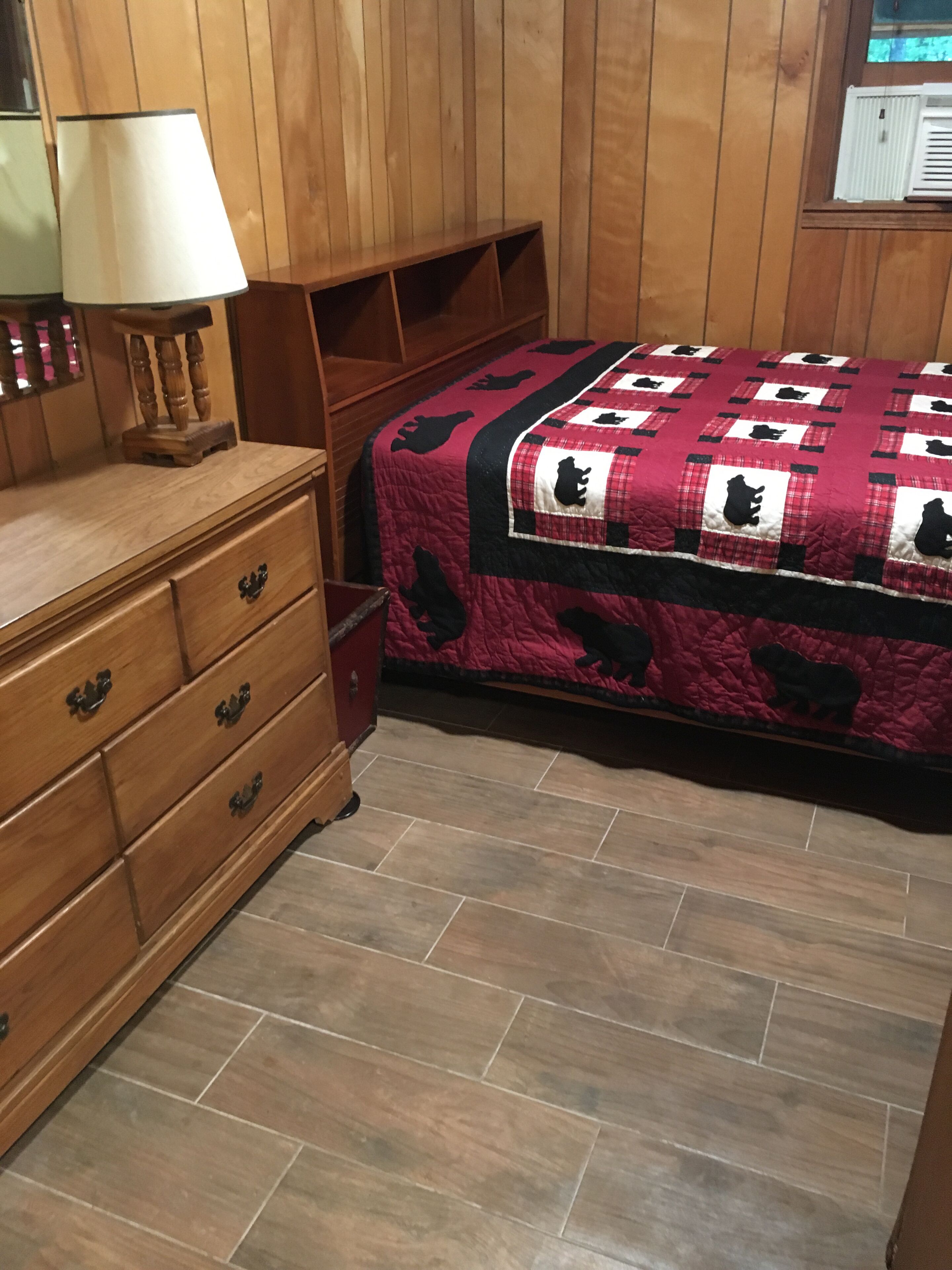 Une chambre à coucher avec une commode en bois, un lit recouvert d’une courtepointe rouge et noire, et une fenêtre avec des stores.