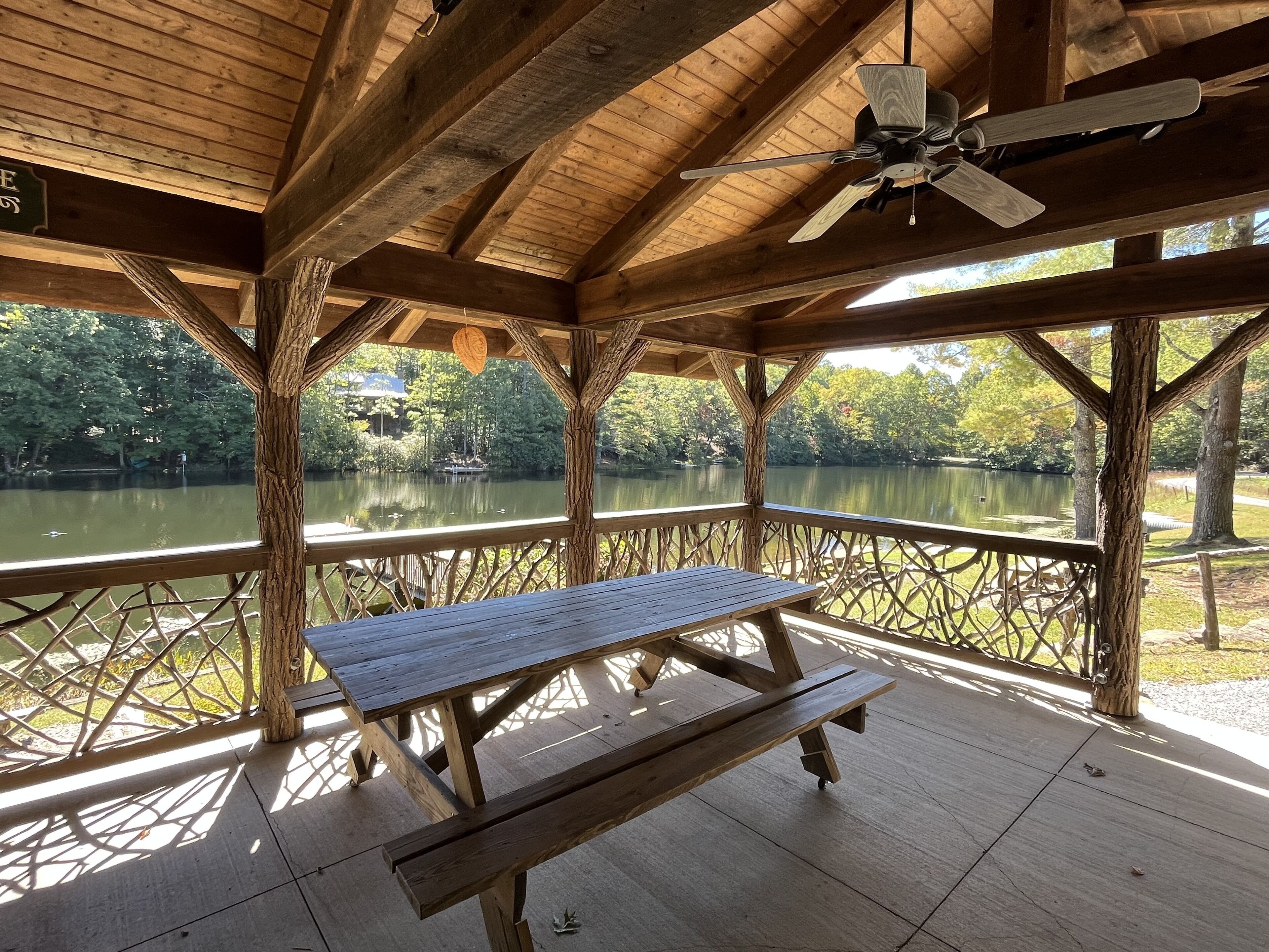 Un pabellón de madera con una mesa y bancos, con vistas a un lago tranquilo con árboles y un muelle al fondo.