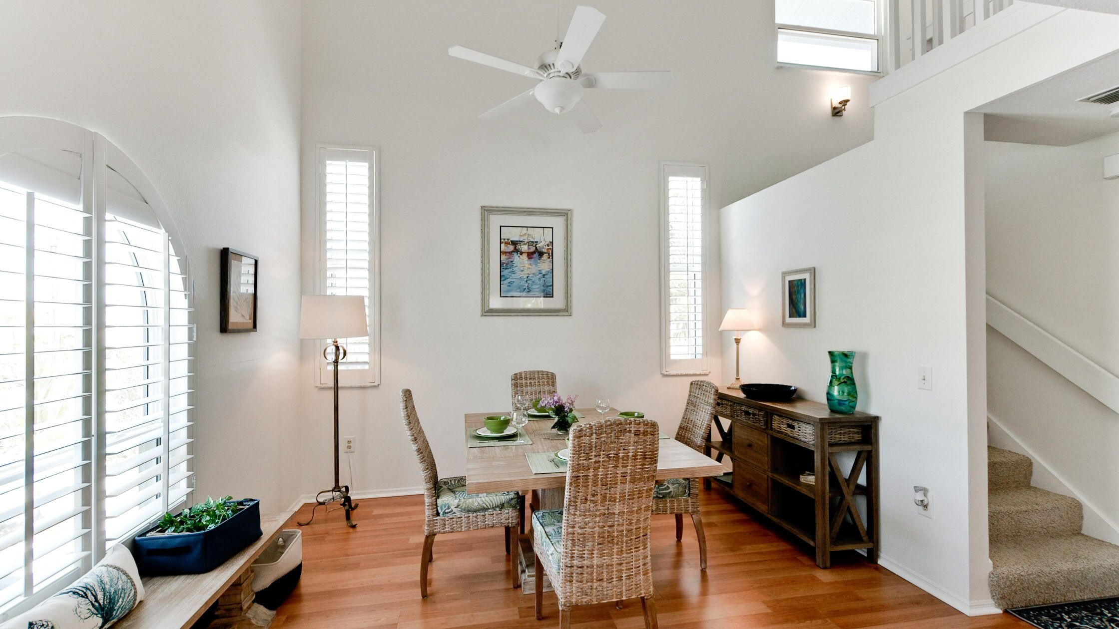 A dining area with a table set for four, surrounded by wicker chairs, a wooden sideboard, and a ceiling fan.