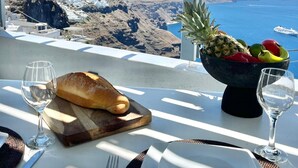 Mesa servida con hogaza de pan, frutera, copas de vino y cubiertos, con vista a un paisaje costero con montañas y el mar.