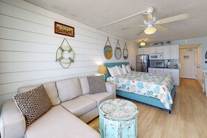 A cozy bedroom with a bed, a sofa, a small round table, and a kitchenette in the background.