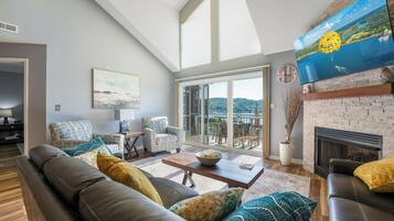 A spacious living room with a sofa, armchairs, a coffee table, and a fireplace. There is a large window with a view of a lake and a painting above the fireplace.