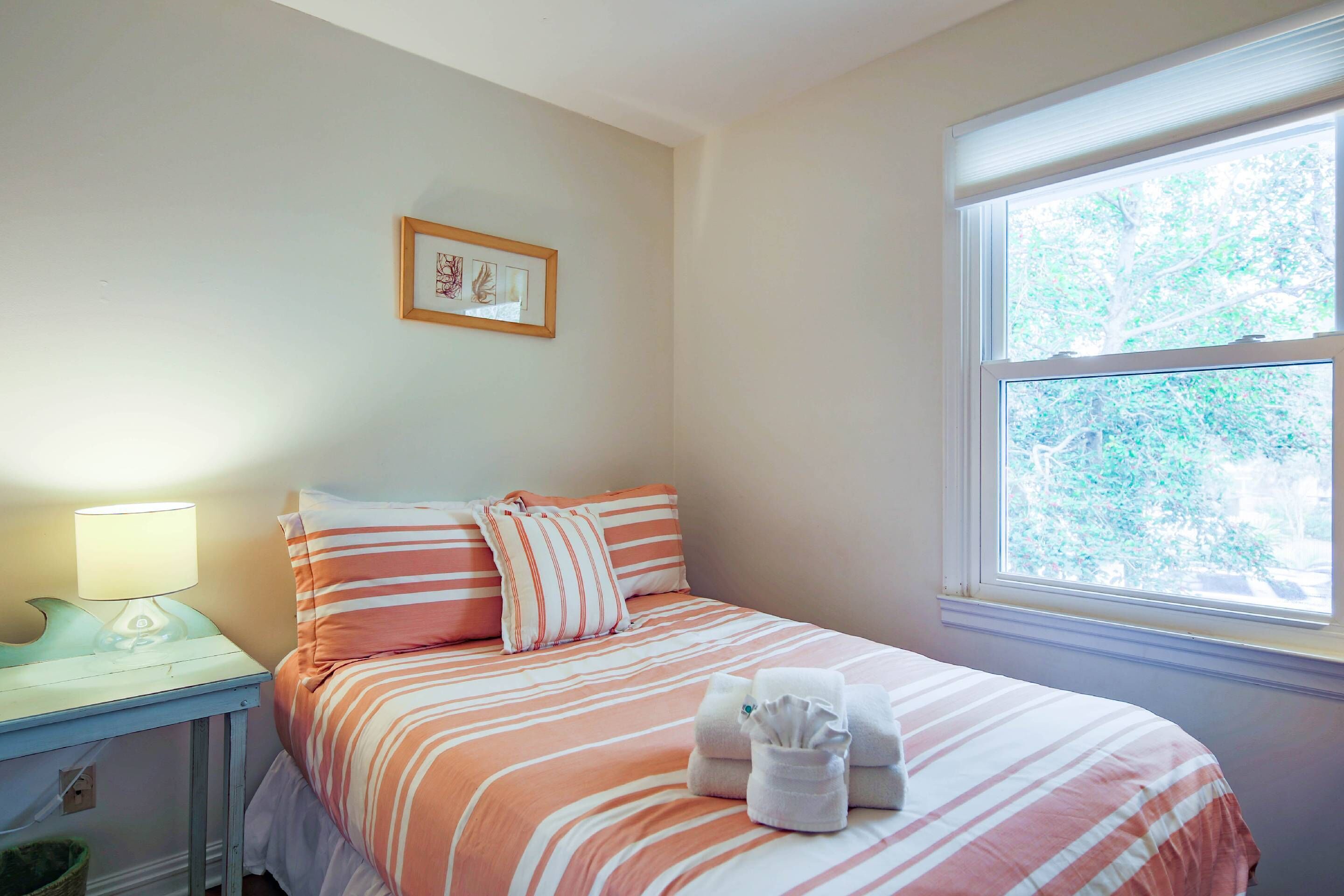 A neatly made bed with striped bedding, a bedside table with a lamp, and a window with a view of trees.
