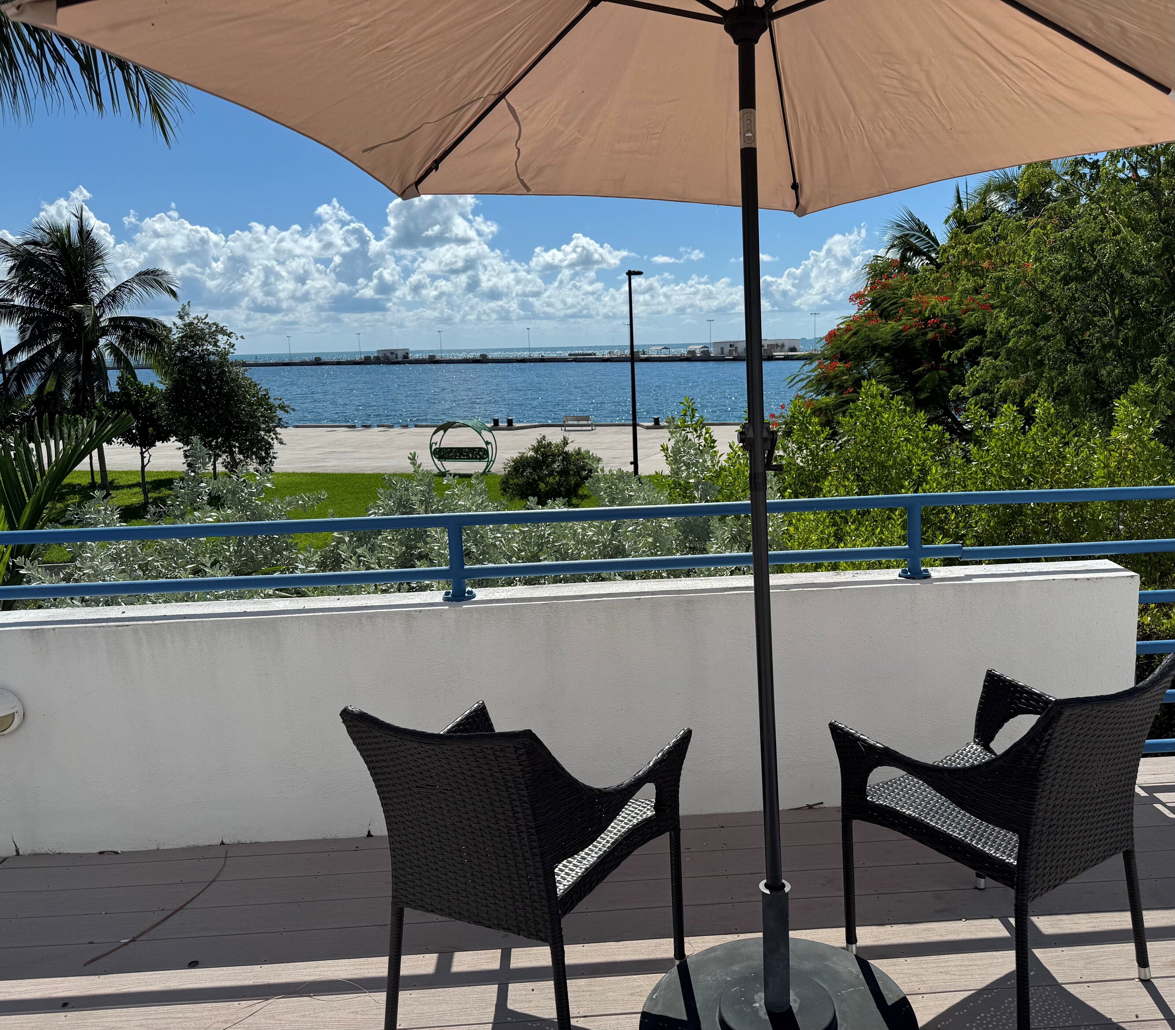 A seaside patio with two wicker chairs and a large umbrella, overlooking a beach and the ocean.