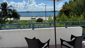 A seaside patio with two wicker chairs and a large umbrella, overlooking a beach and the ocean.