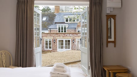 A bedroom with a bed, a wooden nightstand, a mirror, and a view of a brick house through an open door.