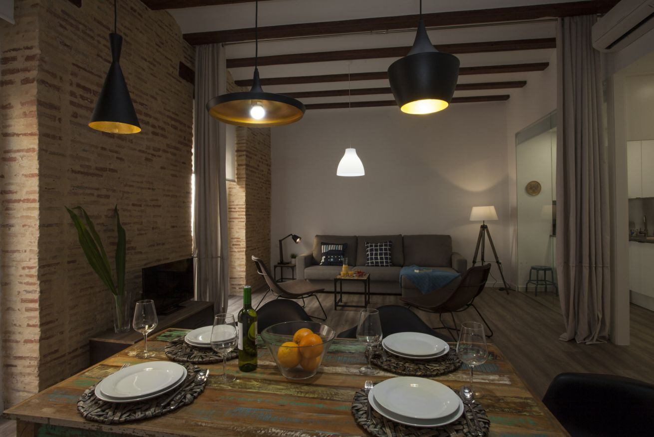 Un coin repas agrémenté d’un mur en briques au style rustique, d’une table en bois dressée pour quatre personnes et de luminaires suspendus.