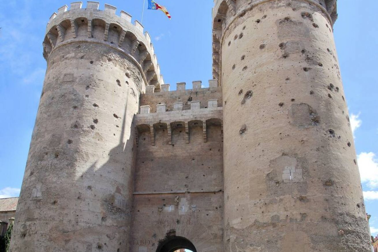 A historic stone castle with two large cylindrical towers, a central gate, and a flag.