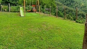 A grassy backyard with a swing set and a slide, surrounded by a stone wall and flowering plants.