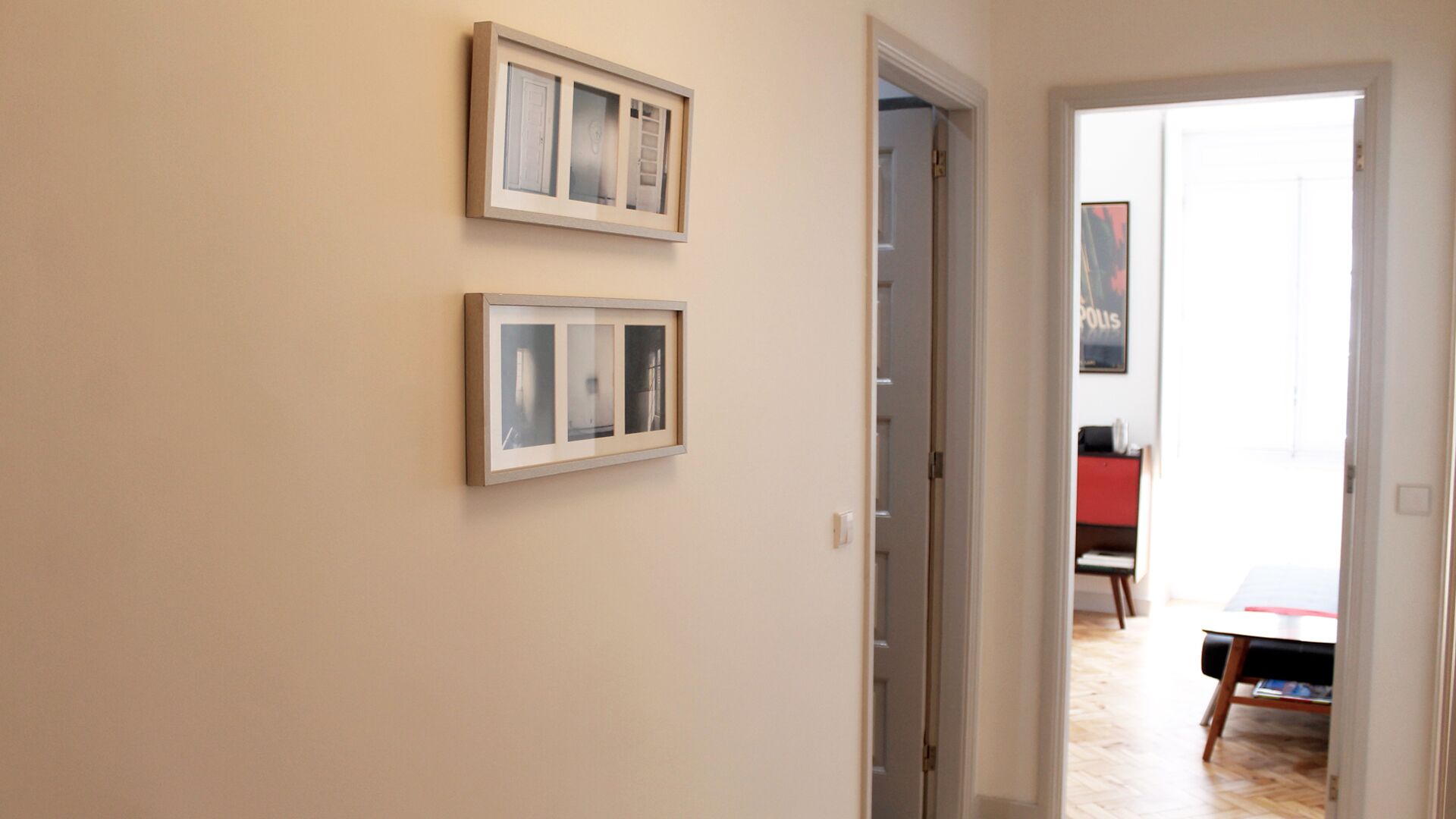A hallway with two framed pictures on the wall, a door leading to a room with a table and chairs, and a poster on the wall.