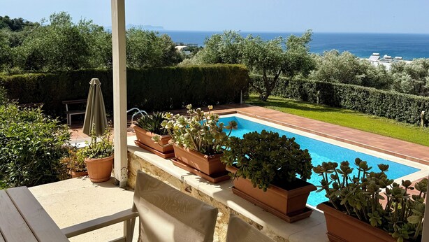 Zona junto a la piscina con vista al mar, plantas en macetas y mobiliario exterior.