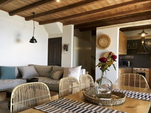 A cozy living room with a wooden ceiling, a sofa with cushions, wicker chairs, a dining table with a vase of flowers, and a kitchen area visible through an open doorway.
