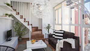 A modern living room with a staircase, a white ceiling light, a brown sofa, and a large window. - Botanico Atico Duplex 302 (Valencia ciudad)
