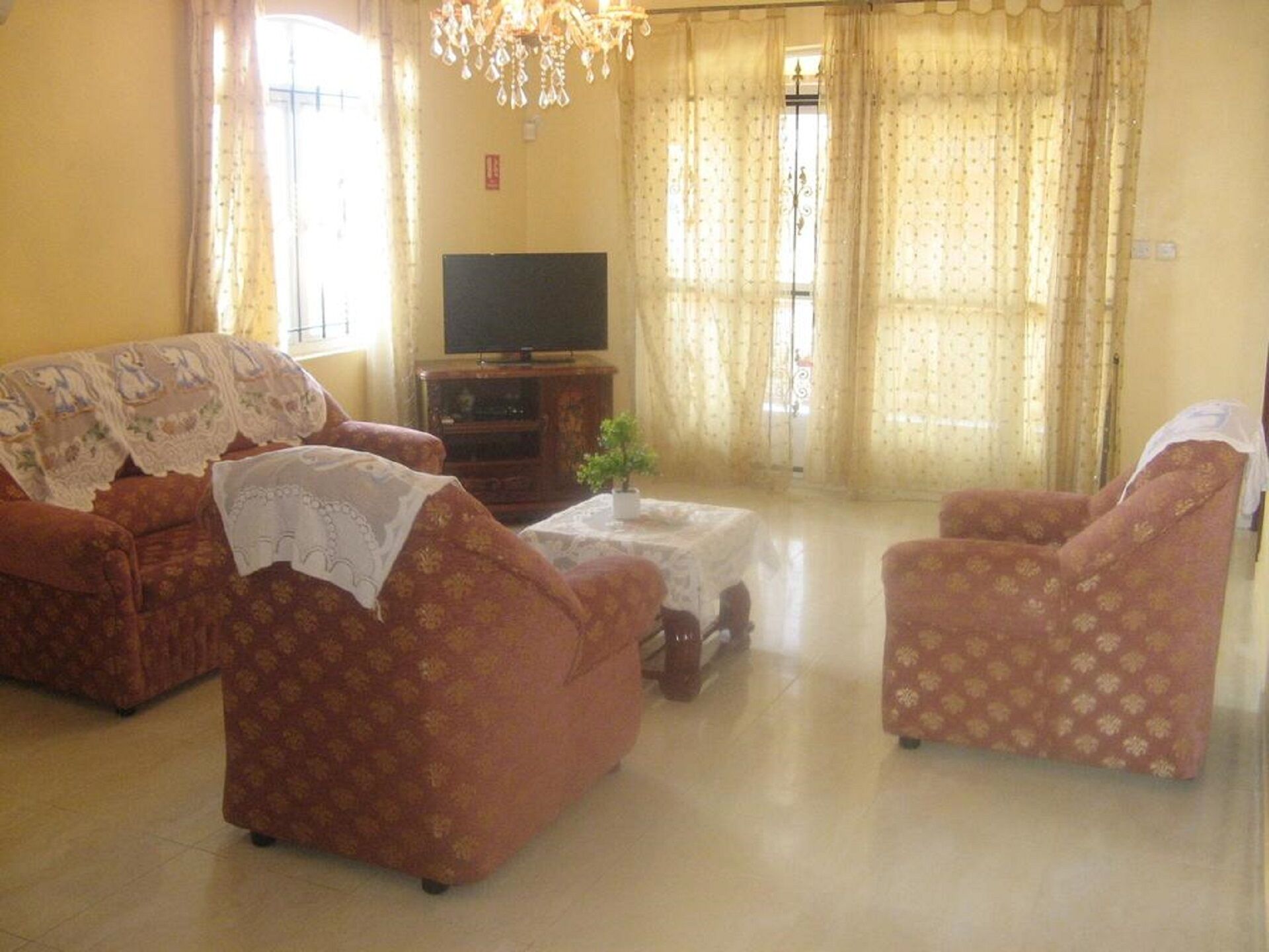 A living room with a sofa, armchair, and a small table. There is a television on a stand and a chandelier hanging from the ceiling.