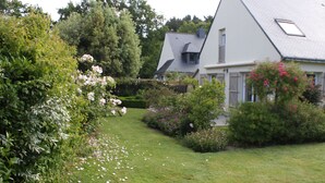 Een huis met een schuin dak, omringd door weelderig groen en bloeiende planten.