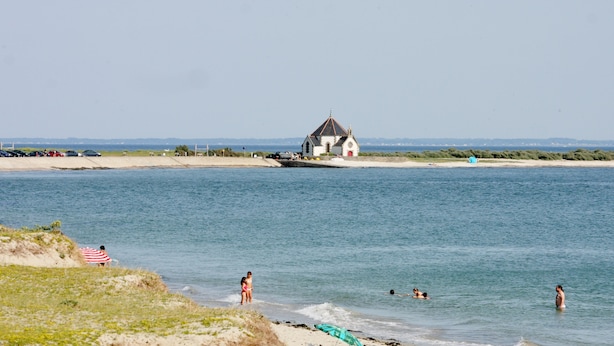 Een strand met een klein gebouw, heldere lucht en mensen die zwemmen en in het water staan.