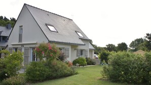 Een huis met twee verdiepingen, een schuin dak, een kleine tuin met struiken en bloemen, en een zichtbare schoorsteen.