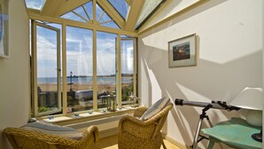 A room with a large bay window overlooking a beach, wicker chairs, a telescope, and a framed picture on the wall.
