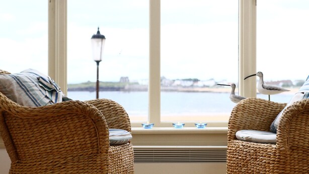 A room with wicker chairs facing a window overlooking a body of water, with a lamp and a bird figurine on the windowsill.