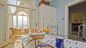 A dining area with a glass table, wicker chairs, a basket of bread, a bowl of oranges, and a blue mug.