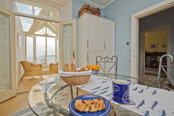A dining area with a glass table, wicker chairs, a basket of bread, a bowl of oranges, and a blue mug.
