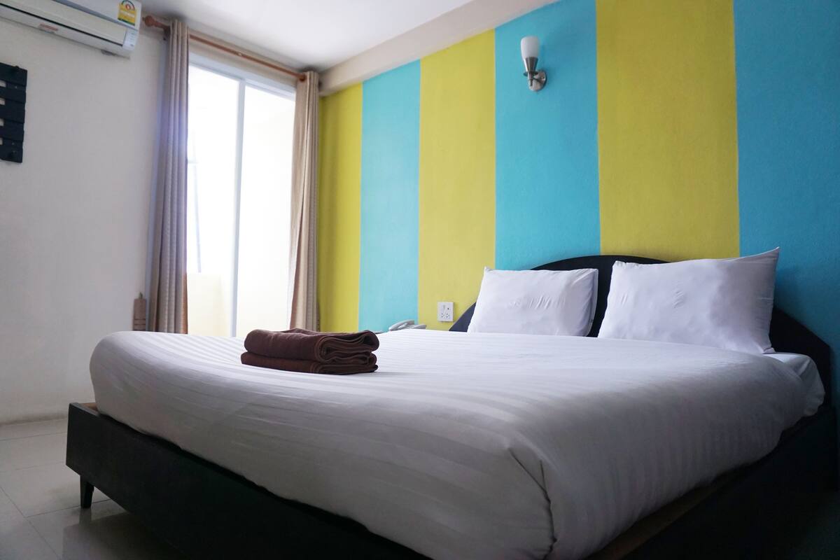 A neatly made bed with white linens and a folded brown towel, a bedside lamp, and a window with curtains.