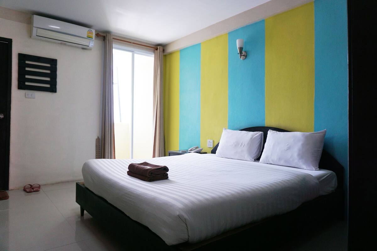 A hotel room with a bed, a nightstand, an air conditioner, and a window with curtains.