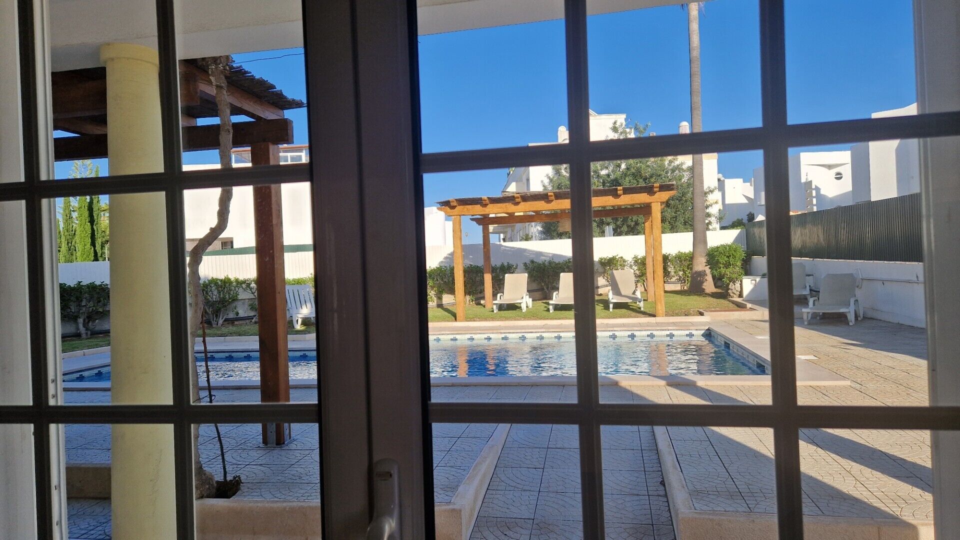 Blick durch ein Fenster auf einen Poolbereich mit einer Pergola, Liegestühlen und blauem Himmel.