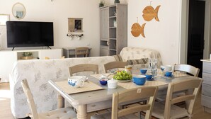 Un coin repas avec une table dressée pour quatre personnes, entourée de chaises. Un téléviseur est fixé au mur, ainsi qu’un meuble avec des étagères et une décoration murale représentant des poissons.