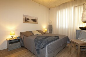 Ein Schlafzimmer mit Bett, Nachttischen, einem Sessel, einer Lampe, einem Gemälde und einem Fenster mit Vorhängen.