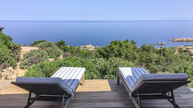 Two sun loungers on a wooden deck overlooking a calm sea with a rocky landscape in the background.
