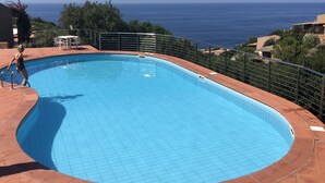 A swimming pool with a view of the sea, surrounded by a metal fence and a patio area with chairs.
