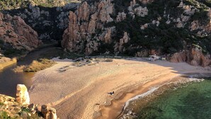 A coastal landscape with a sandy beach, rocky cliffs, and a clear water body.